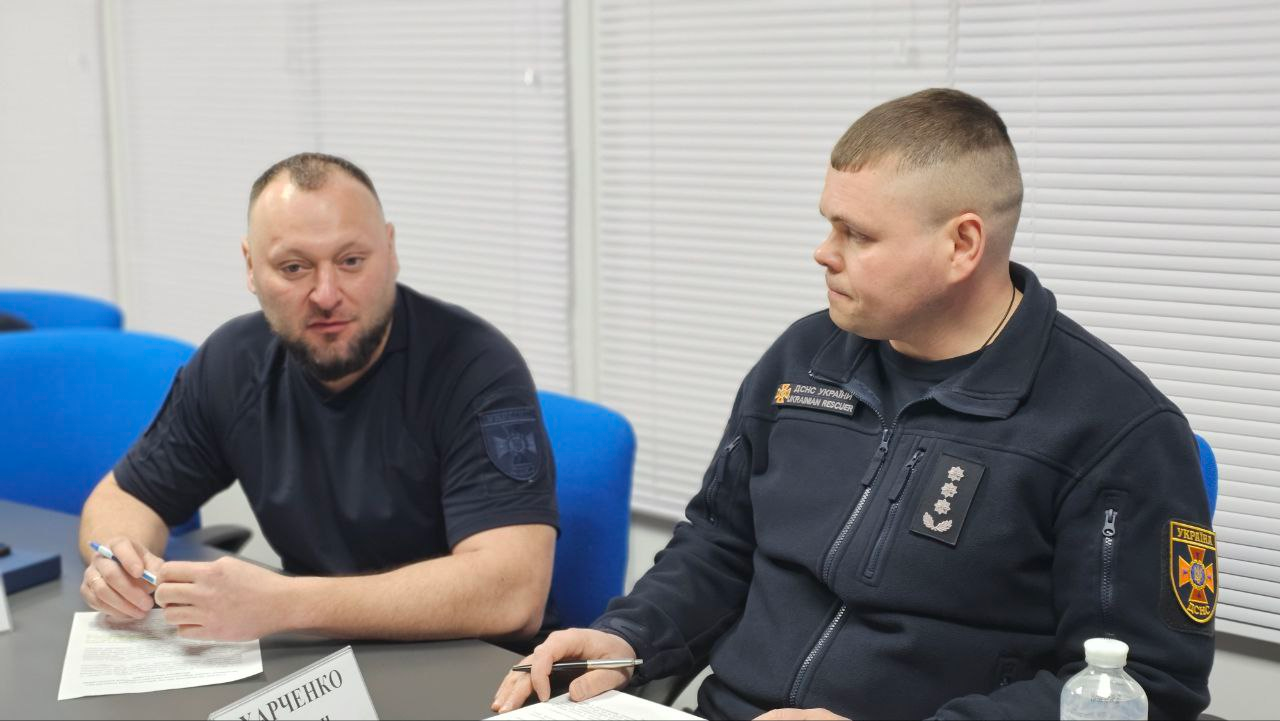 Двоє представників рятувальної служби у формі сидять за столом під час наради. Перед ними лежать документи, ручки та розставлені іменні таблички. Один із чоловіків тримає ручку, інший зосереджено дивиться перед собою.