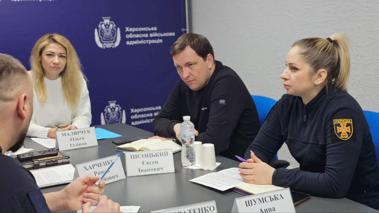 Робоча зустріч за круглим столом: жінка у світлому одязі та чоловік і жінка у темному вбранні (остання — у формі рятувальної служби) слухають співрозмовника. Перед ними стоять іменні таблички та пляшка води на фоні синього інформаційного стенда.