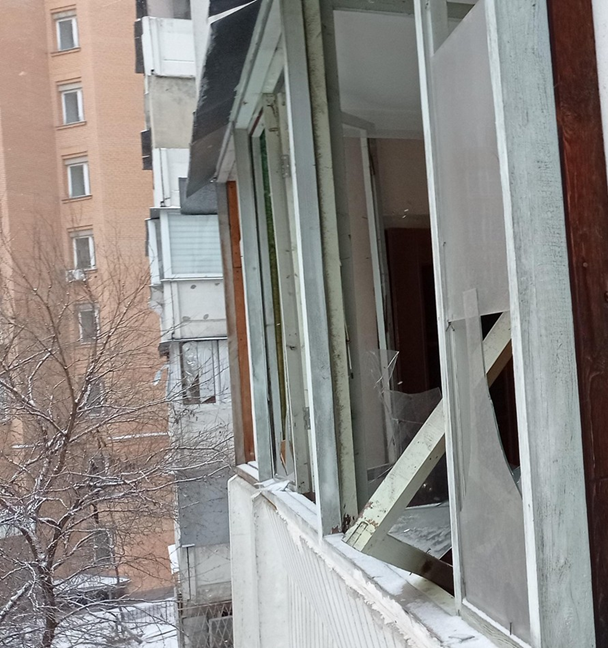 Вигляд зсередини пошкодженого балкона житлового будинку. Віконні рами деформовані, скло вибите, а конструкції тримаються на залишках кріплень. На фоні видно сусідню багатоповерхівку та засніжені дерева.
