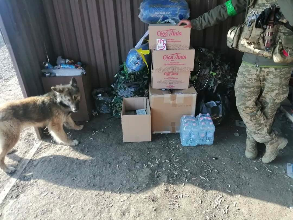Військовий у камуфляжі стоїть поруч із пакунками гуманітарної допомоги, водою та спальними мішками. Біля коробок із написом «Своя лінія» стоїть собака, а на фоні видно маскувальні сітки та речі в приміщенні з металевими стінами.