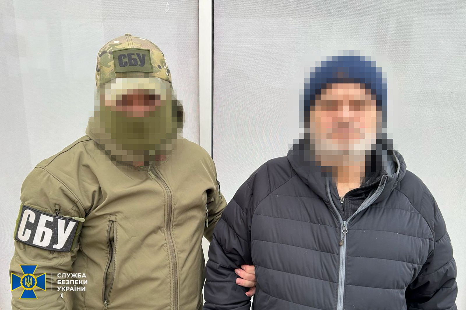 Співробітник СБУ у військовій формі та балаклаві тримає за руку чоловіка у цивільному одязі. Обличчя обох приховані за допомогою пікселізації. У нижньому куті розміщено логотип Служби безпеки України.