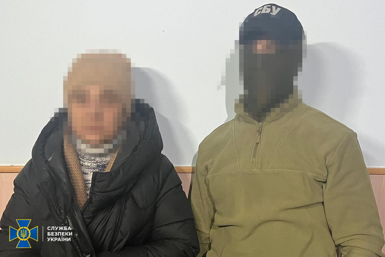 Жінка у зимовому одязі стоїть поруч із представником СБУ, одягненим у тактичне худі та балаклаву. Обличчя фігурантів заретюшовані. У лівому нижньому куті — емблема СБУ.