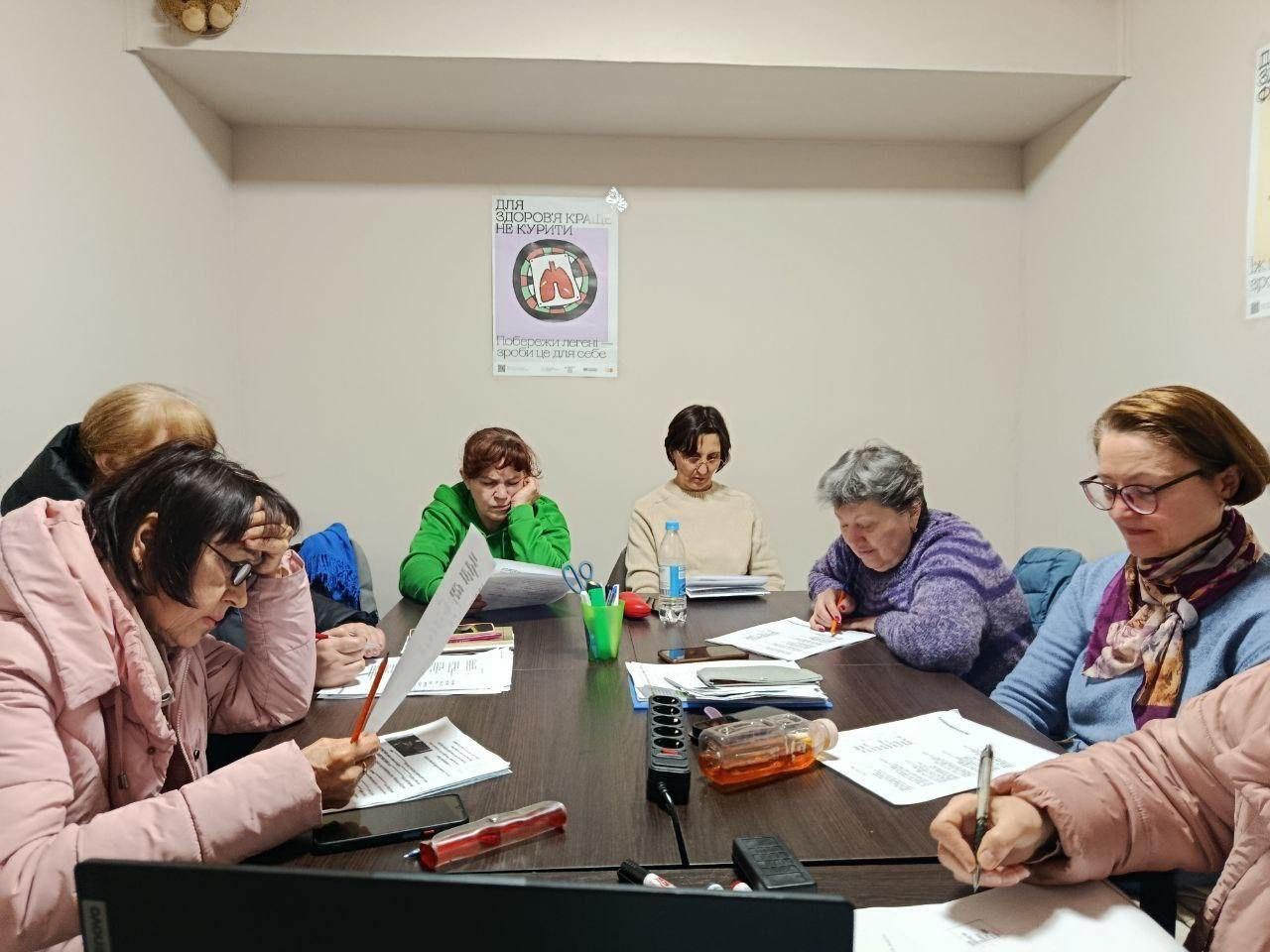 Група жінок сидить за великим столом у приміщенні та працює з паперовими документами. На стіні над ними розміщено плакат про шкоду куріння. На столі лежать робочі матеріали, канцелярія та ноутбук.