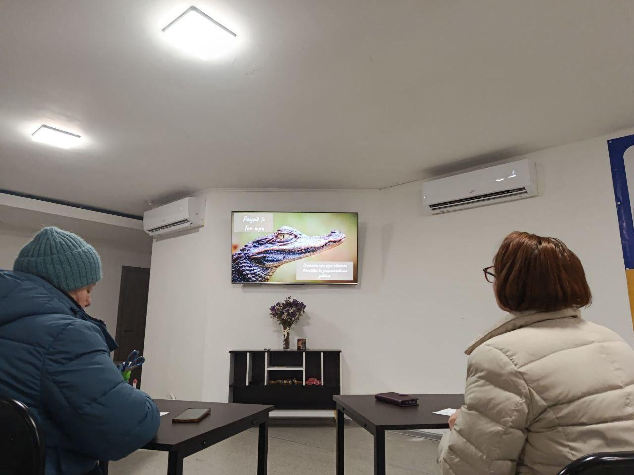 Двоє людей сидять за столами у світлій аудиторії та дивляться на екран телевізора, на якому зображено голову крокодила. У приміщенні працюють кондиціонери, а під екраном стоять квіти у вазі.