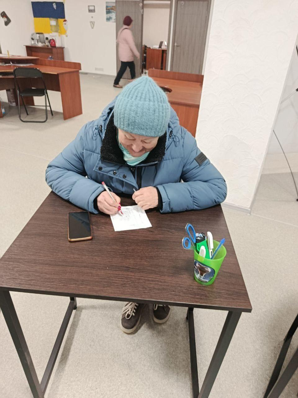 Жінка у блакитній зимовій куртці та шапці сидить за столом і заповнює паперову анкету або документ. Поруч на столі лежить смартфон та стоїть склянка з канцелярським приладдям. На задньому плані видно офісне приміщення з робочими столами.