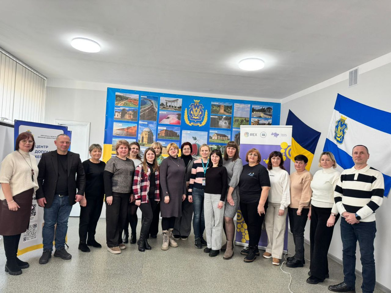 Групове фото учасників заходу у світлому приміщенні. Люди стоять на фоні стенда з фотографіями визначних місць Херсонщини, державного прапора України та прапора Херсонської області. Поруч встановлено інформаційні банери з логотипами організаторів.