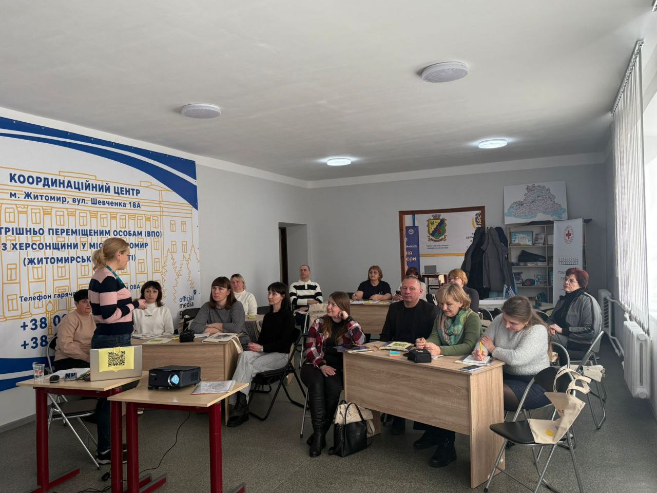 Група людей сидить за робочими столами в аудиторії Координаційного центру та уважно слухає спікерку, яка стоїть біля проєктора. У приміщенні розміщено інформаційні банери, офісну техніку та символіку організацій-партнерів.