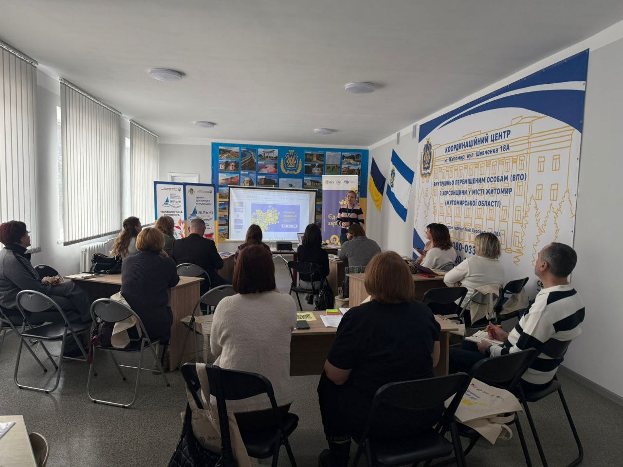 Група людей сидить за столами в аудиторії та спостерігає за презентацією, яку проводить спікерка біля великого екрана. На стіні розміщено банер «Координаційний центр для внутрішньо переміщених осіб з Херсонщини у місті Житомир», а поруч стоять державний прапор України та прапор Херсонської області.