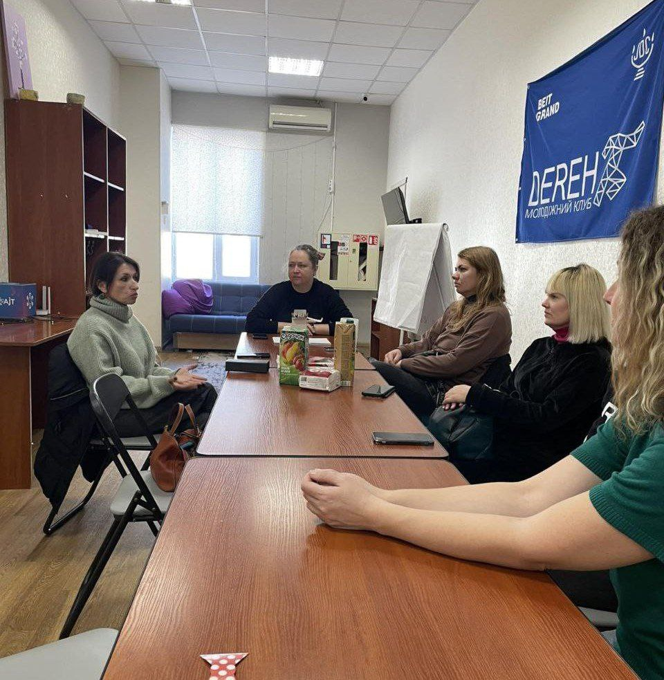 Група жінок проводить робочу зустріч за довгим столом у світлому офісному приміщенні. На стіні розміщено синій прапор із логотипом молодіжного клубу «DEREH». На столі стоять напої та лежать особисті речі учасників.