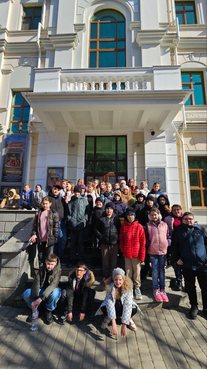 Велика група дітей та підлітків позує для спільного фото на сходах перед входом до великої білої будівлі з колонами та арковими вікнами. Поруч із входом розміщено театральні афіші.