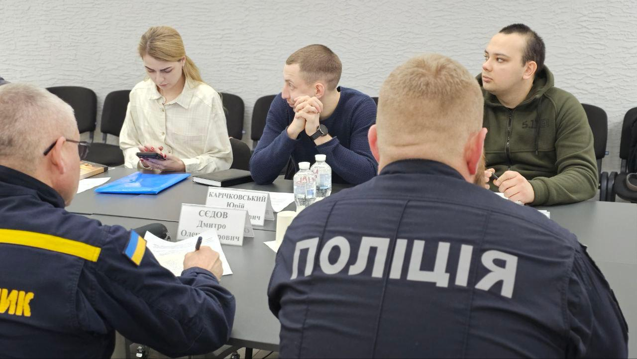 Учасники робочої наради в офіційному приміщенні сидять за столом та обговорюють документи. На передньому плані видно спину представника поліції у формі. На столі розміщені таблички з іменами учасників та пляшки з водою.