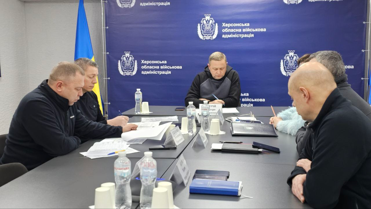 Група людей проводить робочу нараду за довгим столом. Учасники зосереджено вивчають документи та роблять записи. На столі стоять пляшки з водою та паперові стаканчики, а на фоні розташовані державний прапор та синій стенд із офіційною символікою.