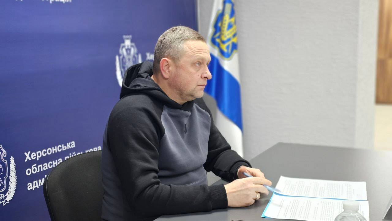 Чоловік у спортивному одязі сидить за робочим столом під час наради. Перед ним лежать документи, а на фоні розташований синій банер Херсонської обласної військової адміністрації та Державний Прапор України.