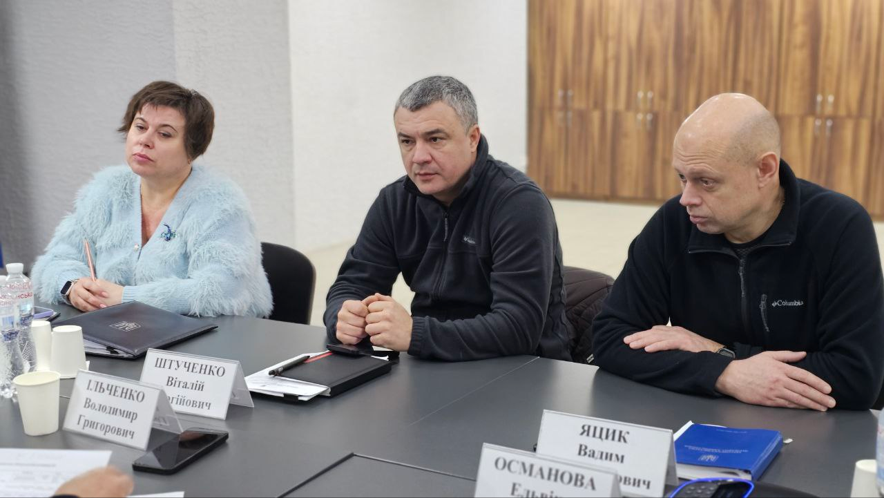 Троє людей — жінка та двоє чоловіків — сидять за столом під час ділової зустрічі. Учасники мають серйозний вигляд і уважно слухають доповідача. Перед ними на столі стоять іменні таблички, паперові стаканчики та лежать документи.