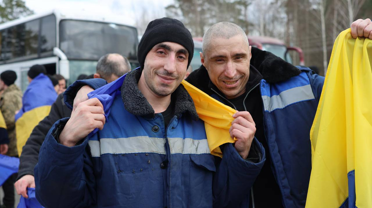 Двоє усміхнених чоловіків у синьому робочому одязі зі світловідбивними смугами обіймаються, тримаючи на плечах державний прапор України. Вони виглядають щасливими на фоні великих пасажирських автобусів та інших людей на відкритому повітрі.