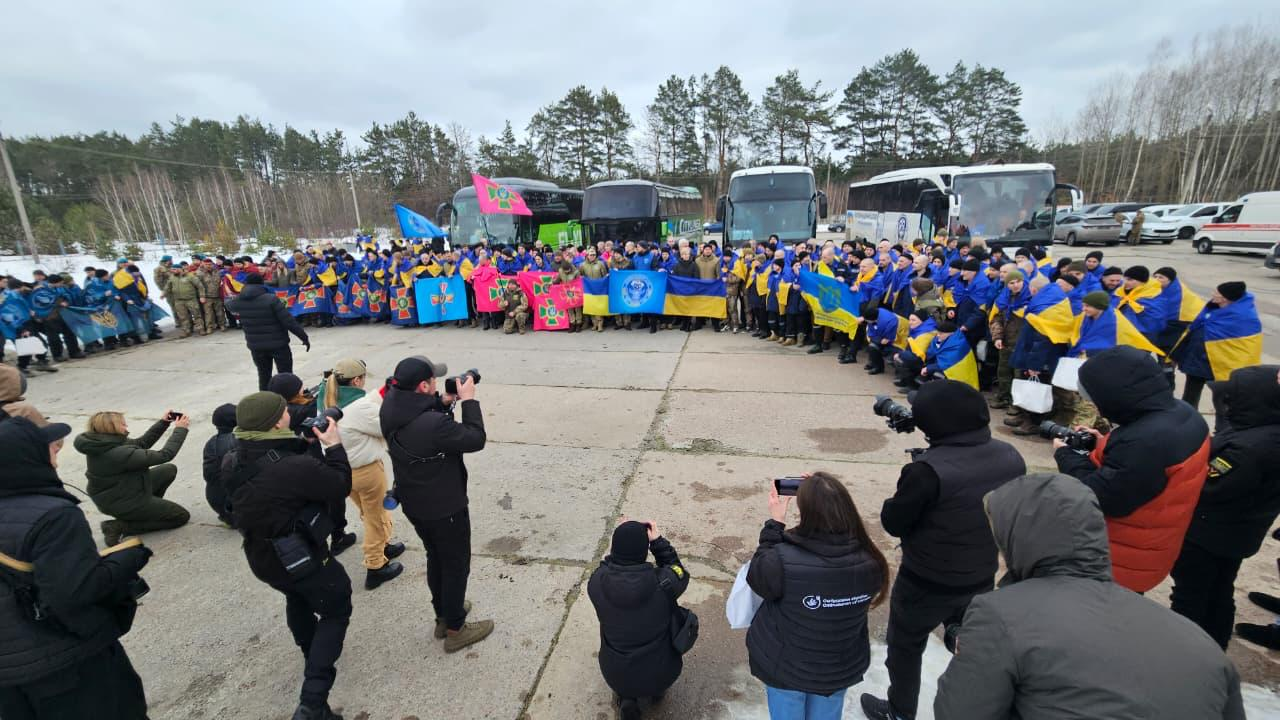 Велика група чоловіків із державними прапорами України та символікою офіційних структур позує для спільного фото на тлі пасажирських автобусів. На передньому плані фотографи та журналісти знімають подію. Дія відбувається на відкритому майданчику біля лісу під похмурим небом.