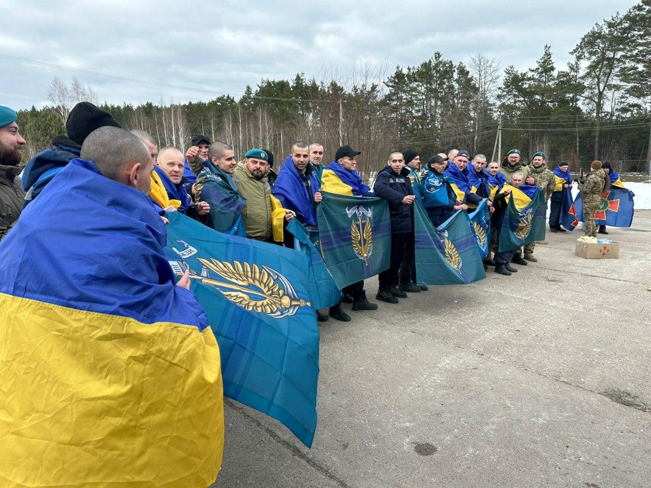 Велика група чоловіків стоїть у шерензі на відкритій місцевості на фоні лісу. Багато з них загорнуті у державні прапори України, а частина тримає великі синьо-зелені стяги з емблемою Координаційного штабу з питань поводження з військовополоненими. Праворуч стоять люди у військовій формі.