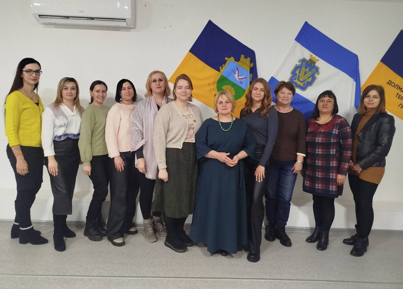Групова фотографія одинадцяти жінок, які стоять у ряд у світлому приміщенні. Вони одягнені у повсякденний та офіційно-діловий одяг. На стіні за ними розміщені три прапори: Державний Прапор України з гербом області, прапор Херсонщини та прапор територіальної громади.