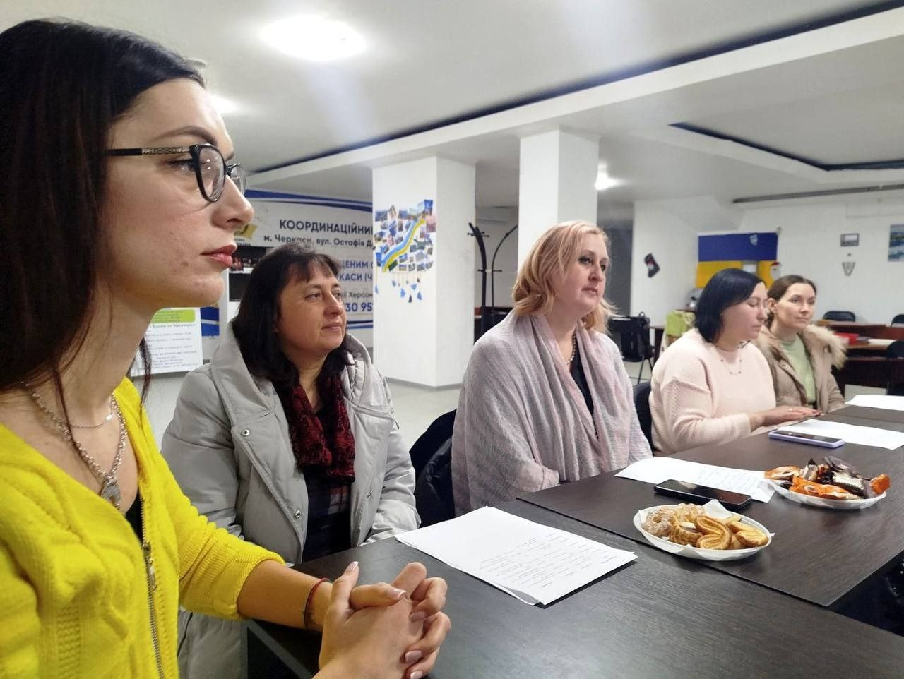 П'ять жінок сидять за довгим столом під час робочої зустрічі чи наради. На столі перед ними лежать документи, смартфони та тарілки з печивом. На задньому плані видно інформаційний стенд із написом «Координаційний» та невеликий прапор України на стіні.