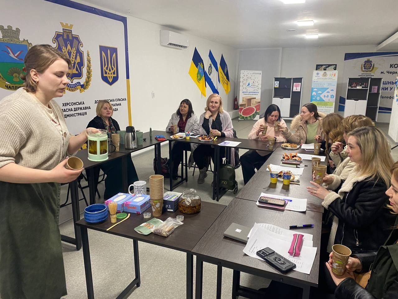 Група жінок проводить неформальну зустріч за столом у світлому приміщенні. На передньому плані жінка готує напої, а інші учасниці спілкуються, тримаючи паперові стаканчики. На столах лежать документи та частування, а на стінах розміщено офіційну символіку та прапори України.