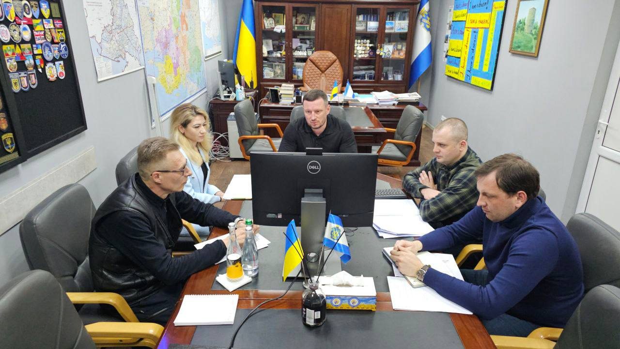 Група людей проводить робочу зустріч у кабінеті за столом, на якому розміщені документи, монітор комп'ютера та маленькі прапорці України. На стінах кабінету видно мапи, стенди та державну символіку.