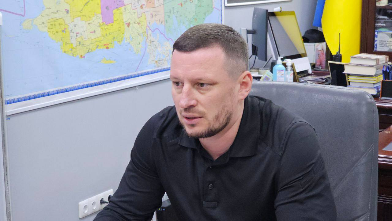 Чоловік у чорній сорочці-поло сидить у робочому кабінеті на фоні настінної мапи. Поруч на робочому столі видно монітор, книги та державний прапор України.