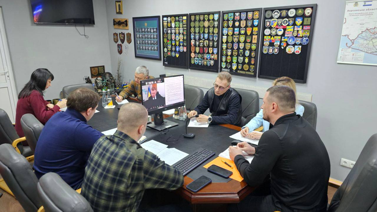Група людей проводить нараду за великим столом, використовуючи відеозв'язок на моніторі. На стіні кабінету розміщені численні стенди з військовими шевронами та мапа Херсонської області.