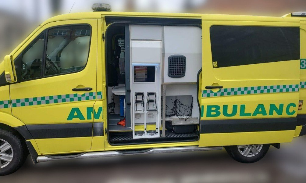 Жовтий автомобіль швидкої допомоги з відчиненими боковими дверима, через які видно спеціальне медичне обладнання та стелажі всередині. На кузові нанесено напис «AMBULANCE» та зелену світловідбивну стрічку.