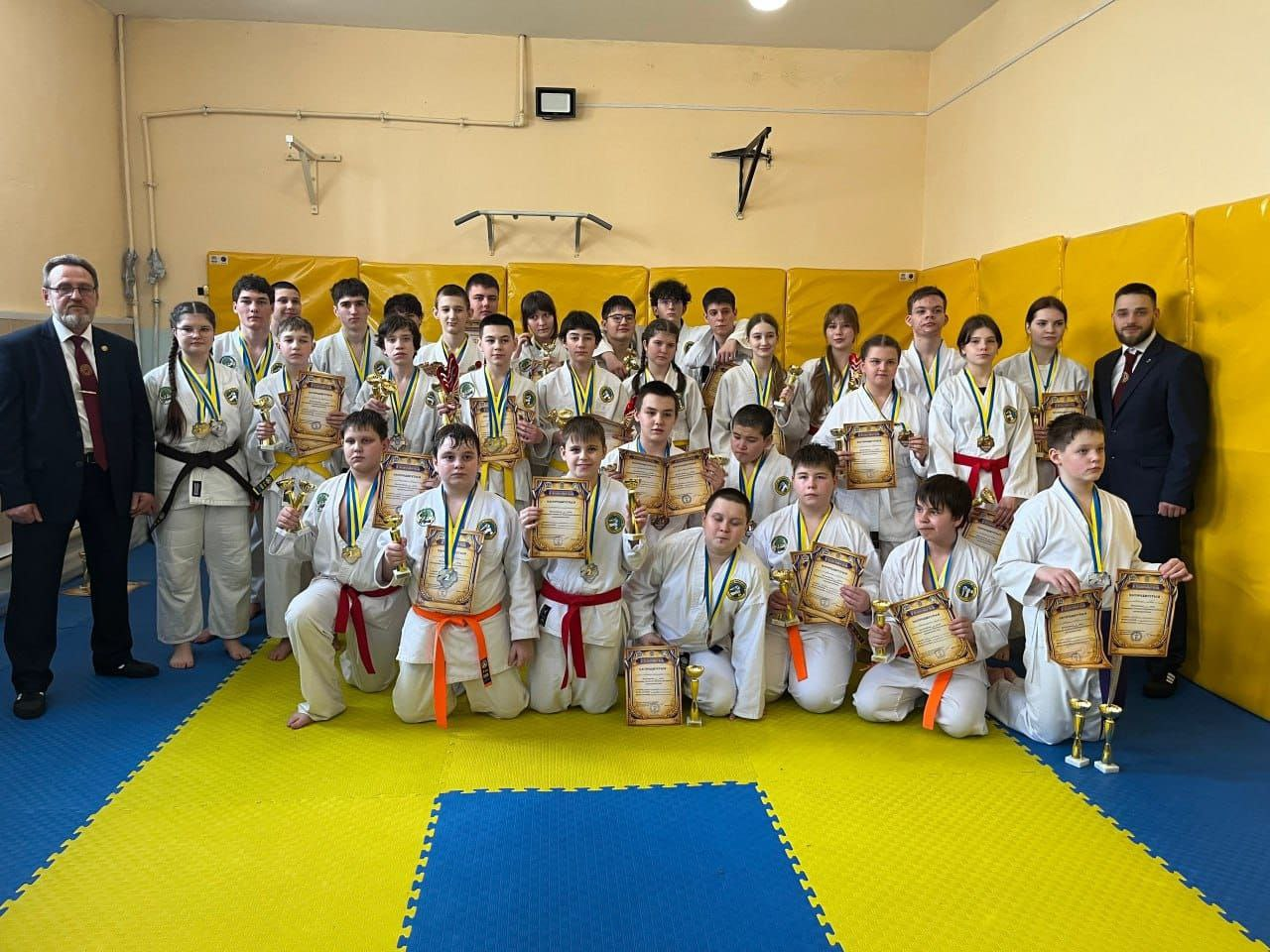 Групове фото юних спортсменів у кімоно після змагань з бойових мистецтв. Діти та підлітки тримають у руках грамоти, кубки та медалі, позуючи на спортивних матах у залі разом із тренерами.