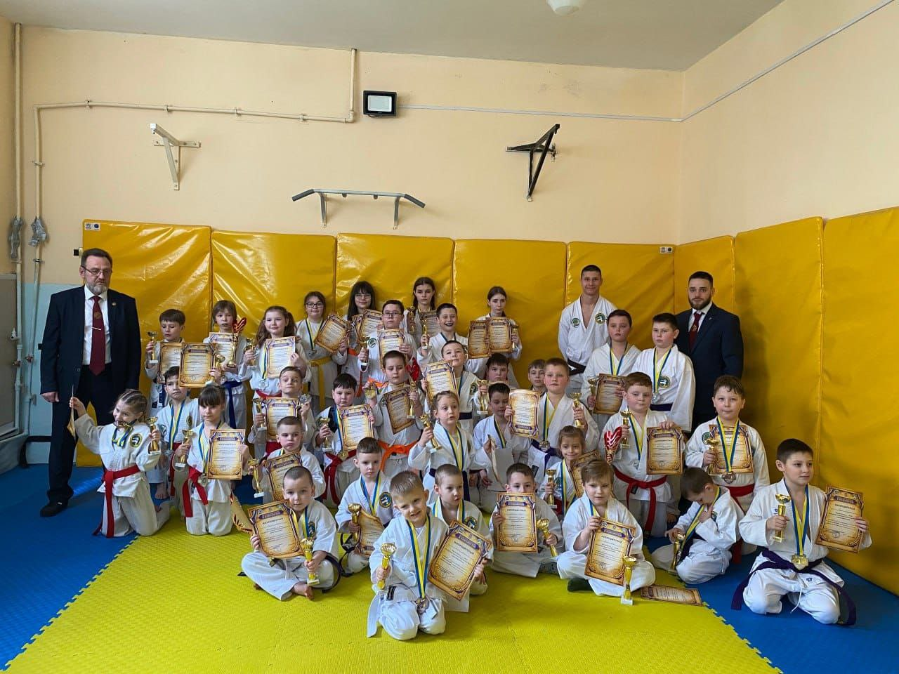 Групове фото дітей у білих кімоно з медалями, кубками та грамотами в руках. Юні спортсмени позують разом із тренерами на жовто-блакитному татамі в спортивній залі після змагань.