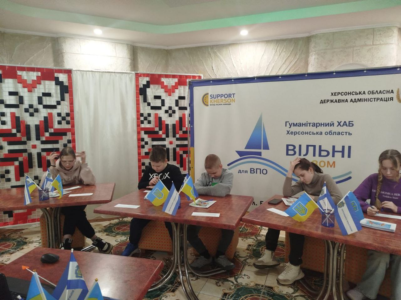 Група дітей та підлітків займається за столами з українською символікою в гуманітарному хабі. Позаду них — банер Херсонської ОДА та декор із традиційним орнаментом вишивки.