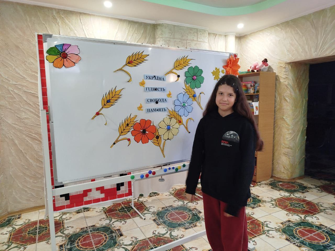 Дівчина стоїть біля білої дошки, прикрашеної паперовими квітами, колоссям та написами про Україну, гідність і свободу. Вона одягнена у темне худі та червоні штани, а подія відбувається у світлій кімнаті з візерунчастою підлогою.