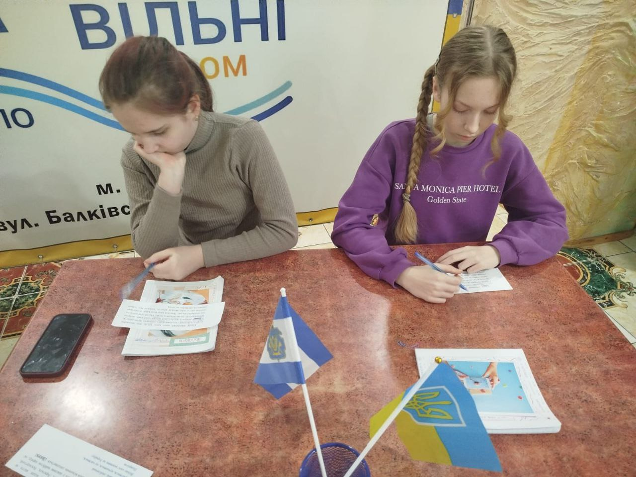 Дві дівчини сидять за столом, зосереджено працюючи з паперами. На столі стоять маленькі прапорці України, а позаду — інформаційний банер гуманітарного центру.