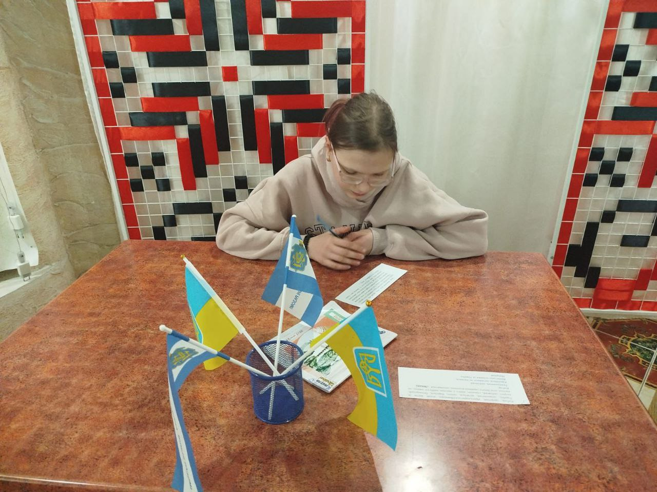 Дівчина в окулярах зосереджено читає роздрукований текст, сидячи за столом. Перед нею стоїть підставка з маленькими прапорцями України, а на фоні — декоративне панно з традиційним червоно-чорним орнаментом вишивки.