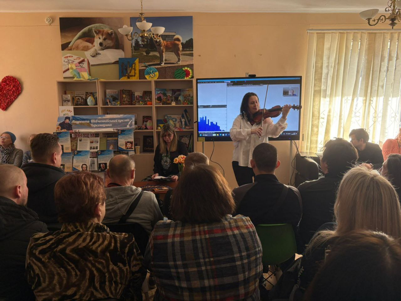 Жінка грає на скрипці перед аудиторією в затишній залі. Позаду неї стоїть великий екран, а збоку розташований стелаж із книгами та інформаційним стендом. Глядачі сидять спиною до камери, спостерігаючи за виступом.