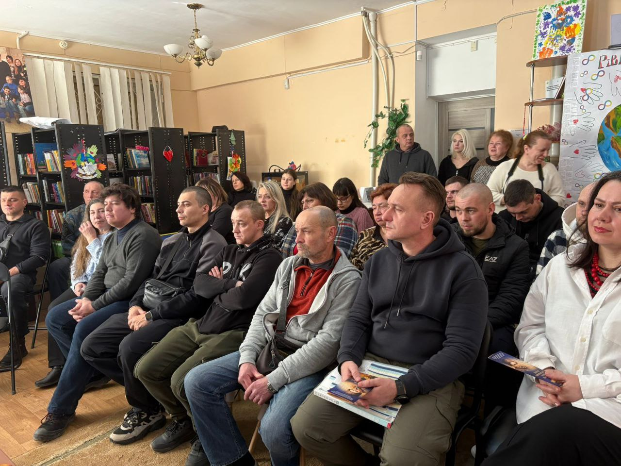 Група людей різного віку сидить у світлій залі під час заходу. Вони зосереджено слухають спікера або дивляться виступ. На задньому плані видно книжкові полиці та плакати на стінах.