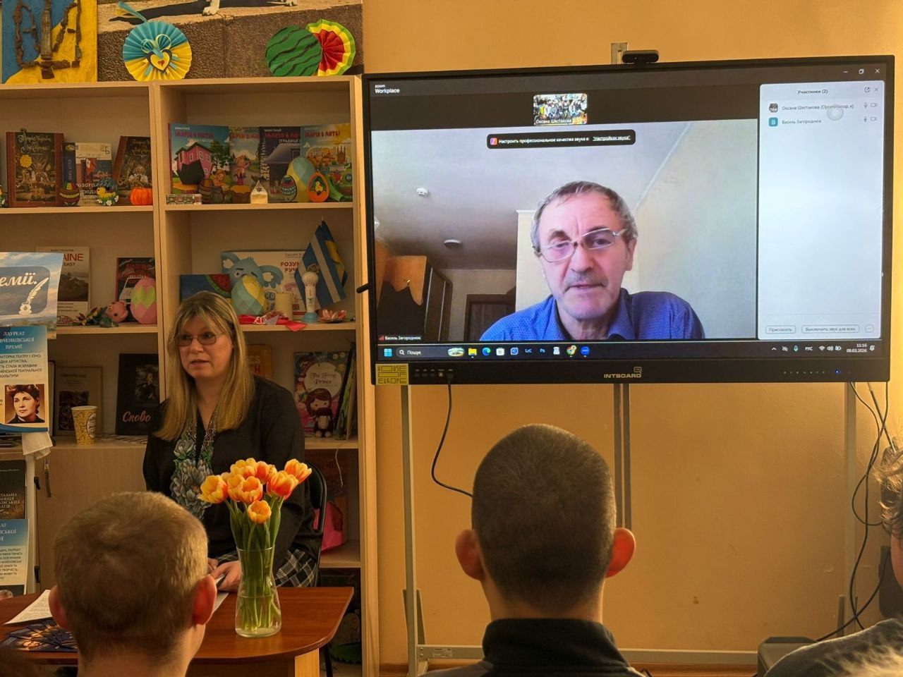 Онлайн-зустріч у приміщенні з книжковими полицями. Жінка сидить за столом із тюльпанами біля великого екрана, на якому транслюється відеодзвінок зі спікером. На передньому плані — слухачі, що спостерігають за подією.