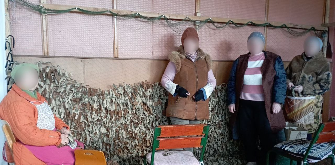 Чотири жінки (обличчя приховані) займаються плетінням маскувальної сітки у приміщенні. Одна жінка сидить ліворуч у помаранчевій куртці, троє інших стоять поруч із великою сіткою, закріпленою на дерев’яній рамі. На задньому плані видно стіну, обшиту панелями.