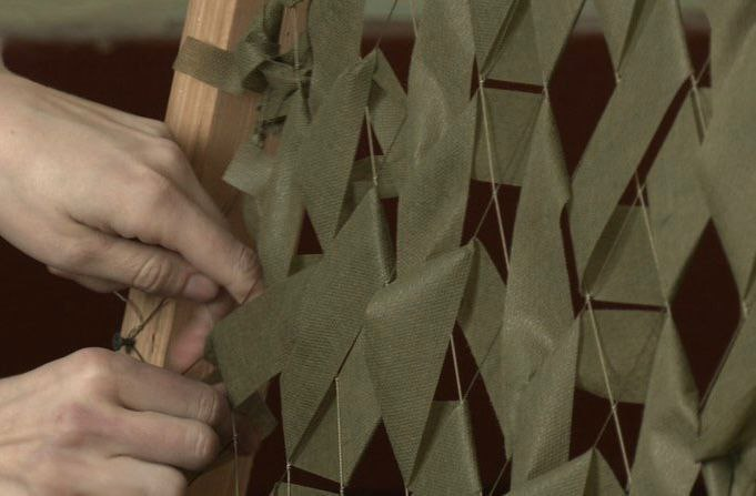 На великому плані зображено руки людини, які вплітають смужки тканини захисного кольору в основу маскувальної сітки, закріплену на дерев'яному каркасі.