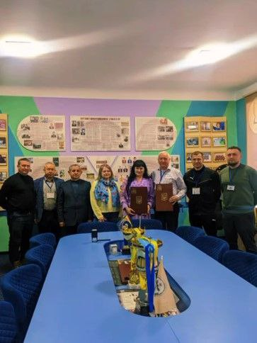 Група людей стоїть у приміщенні біля великого синього столу для нарад. На задньому плані — стіна з інформаційними стендами. Двоє людей у центрі тримають у руках папки з гербом України, що може свідчити про підписання офіційного документа чи меморандуму.