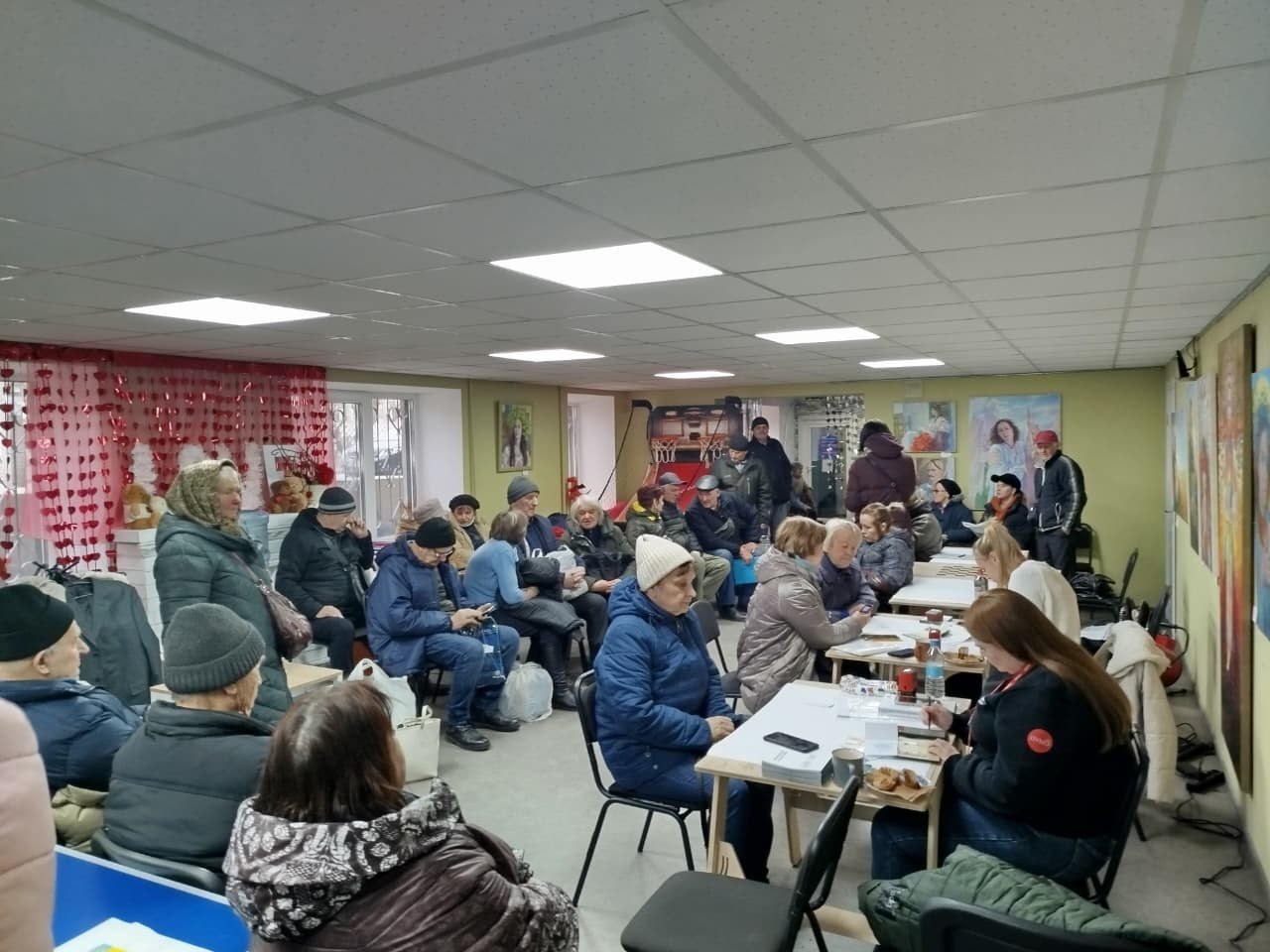 Група людей різного віку в теплому верхньому одязі перебуває у світлому приміщенні. Частина людей сидять у черзі вздовж стін, інші — за столами, де спілкуються з адміністраторами або волонтерами. На стінах видно картини та вікна, декоровані червоними прикрасами.