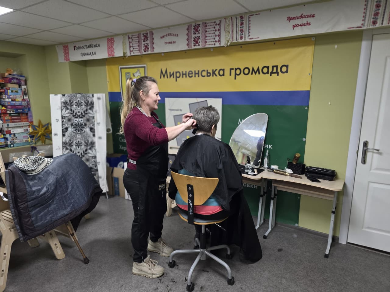Жінка-перукар у чорному фартуху робить стрижку іншій людині в облаштованому приміщенні. На стіні за ними розміщено банер із написом «Мирненська громада», а поруч — стіл із дзеркалом та перукарським приладдям.