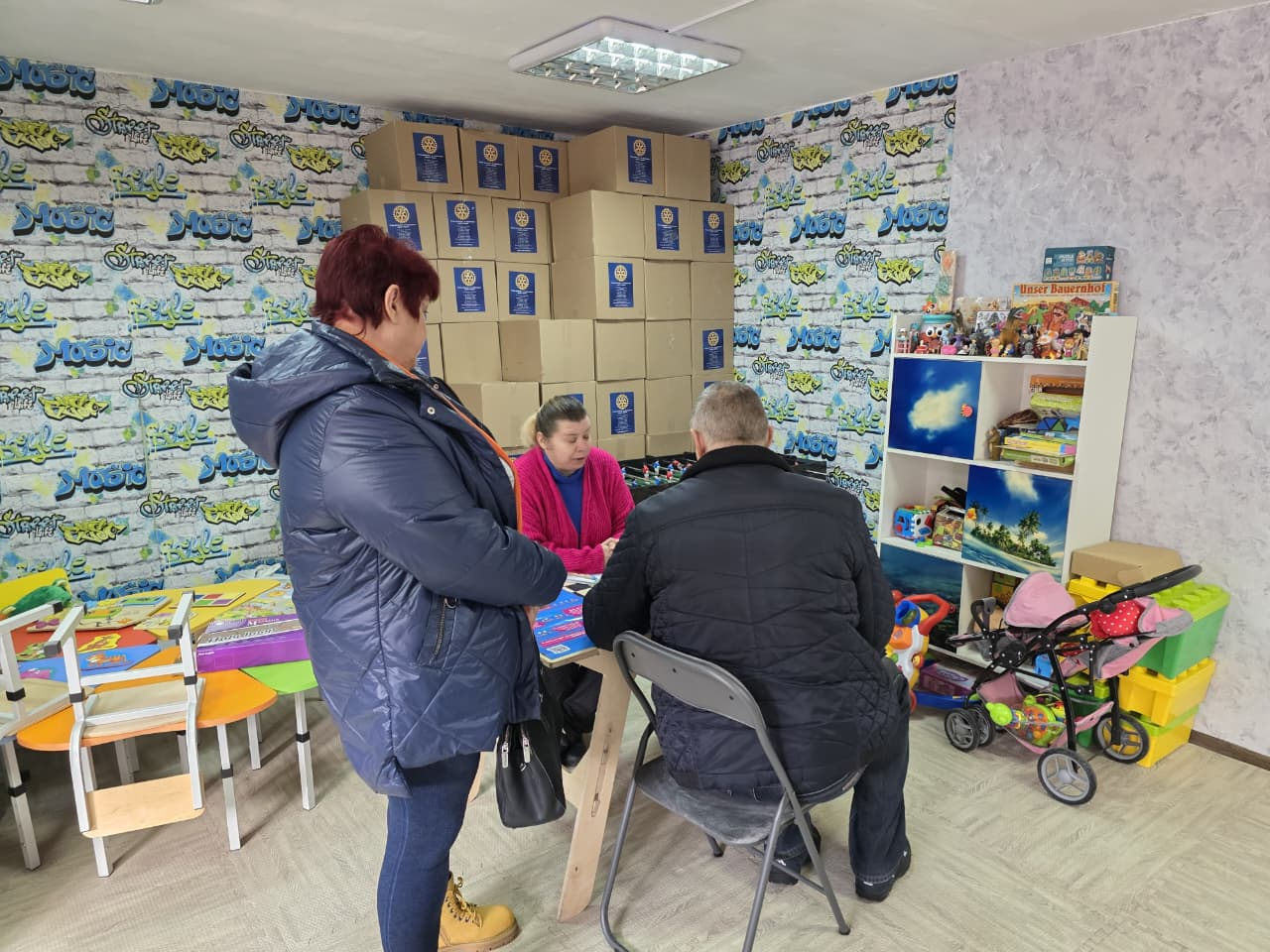 Троє людей спілкуються в приміщенні, облаштованому як пункт допомоги або дитячий простір. На задньому плані складено багато картонних коробок із логотипами, а поруч стоїть полиця з іграшками та дитячий візок.