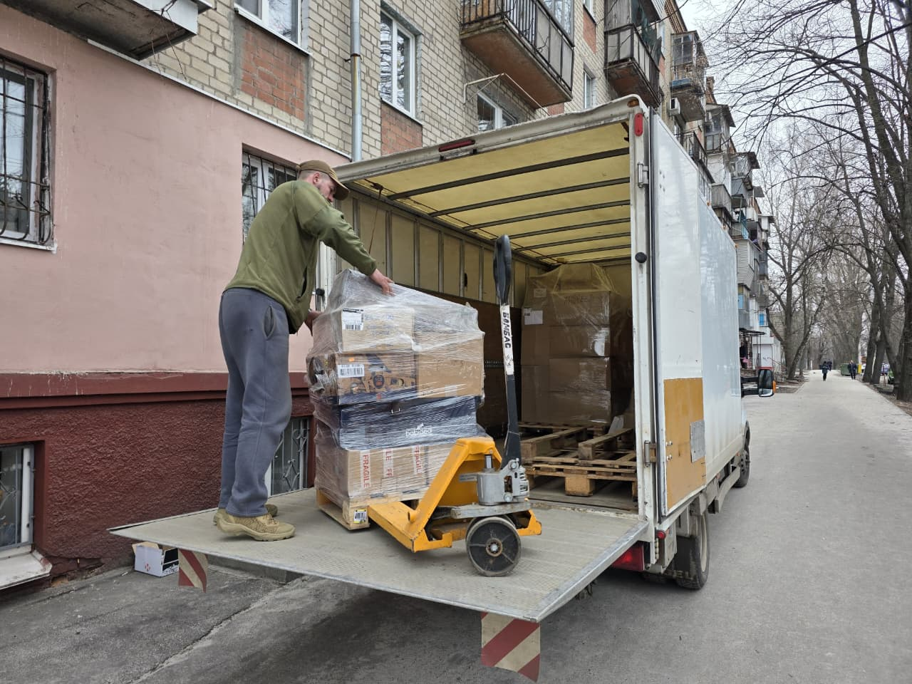 Чоловік на гідроборті білої вантажівки розвантажує піддон із коробками, загорнутими в плівку, за допомогою гідравлічного візка (рокли). Автомобіль стоїть на вулиці біля багатоповерхового житлового будинку.