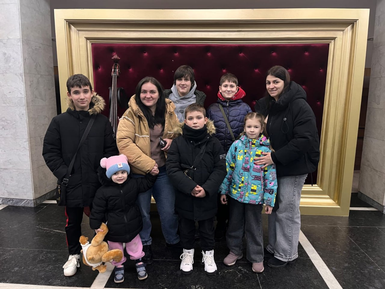 Група дітей та дорослих у верхньому зимовому одязі позує для спільного фото в приміщенні. Вони стоять на фоні великої золотої рами з темно-червоною м'якою оббивкою всередині, що нагадує декоративну фотозону.