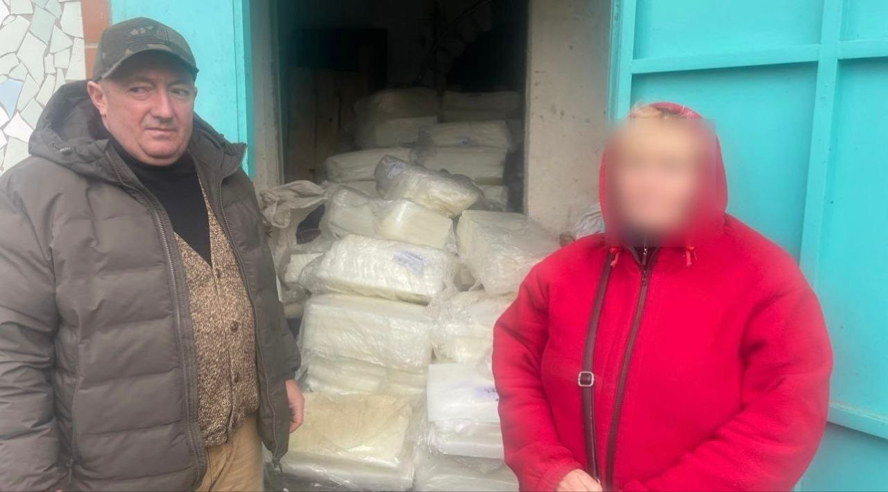 Чоловік у кепці та людина в червоній куртці стоять біля входу до приміщення, що вщент заповнене білими пакунками. Обличчя особи праворуч розмите.