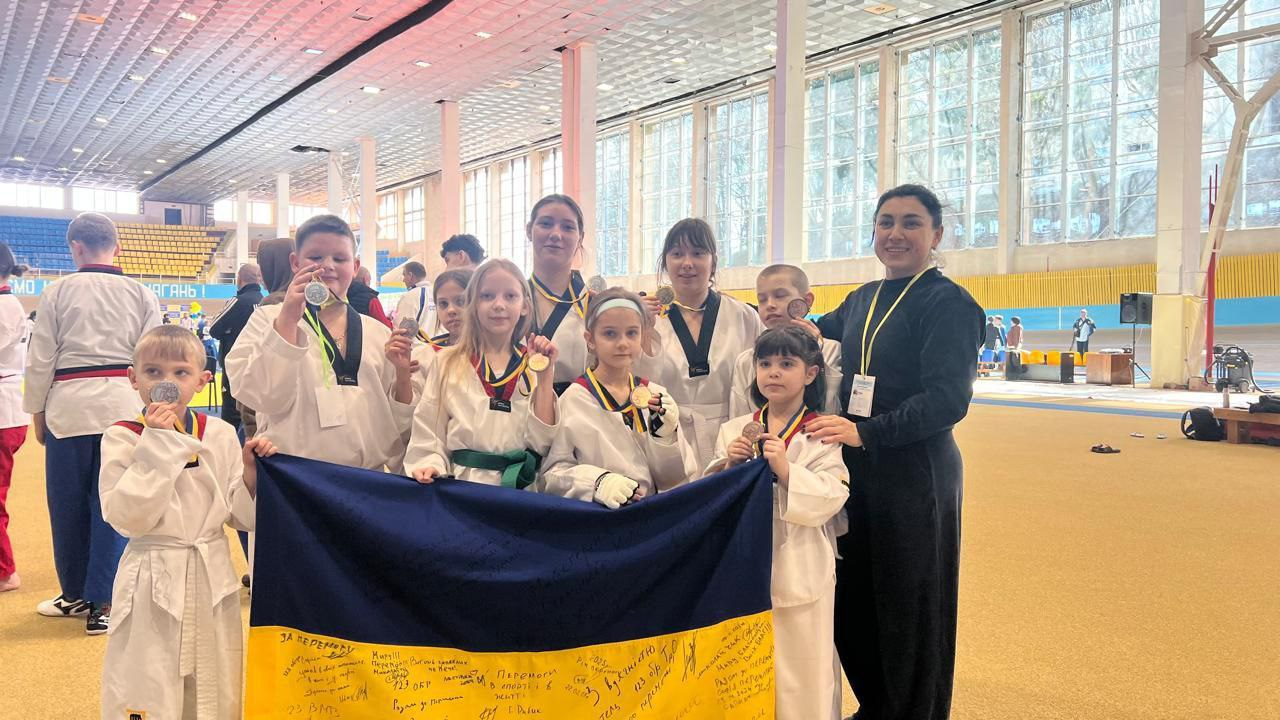 Група юних спортсменів у білих кімоно та їхня тренерка позують у великому спортивному залі. Діти тримають у руках завойовані медалі та розгорнутий український прапор із побажаннями та підписами.