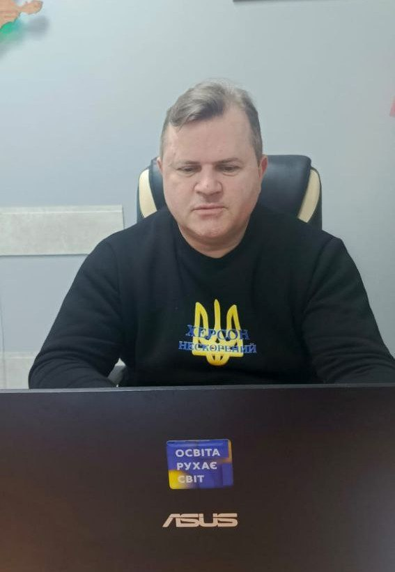Чоловік у чорному світшоті з гербом України та написом «Херсон нескорений» працює за ноутбуком. На кришці комп'ютера розміщено стікер із гаслом «Освіта рухає світ».