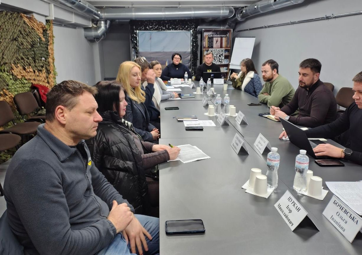 Група людей у цивільному одязі сидить за довгим Т-подібним столом під час наради або зустрічі в приміщенні, схожому на укриття. На столі перед ними розміщені таблички з іменами, пляшки води, папери для записів та мобільні телефони. На стіні ліворуч висить маскувальна сітка.