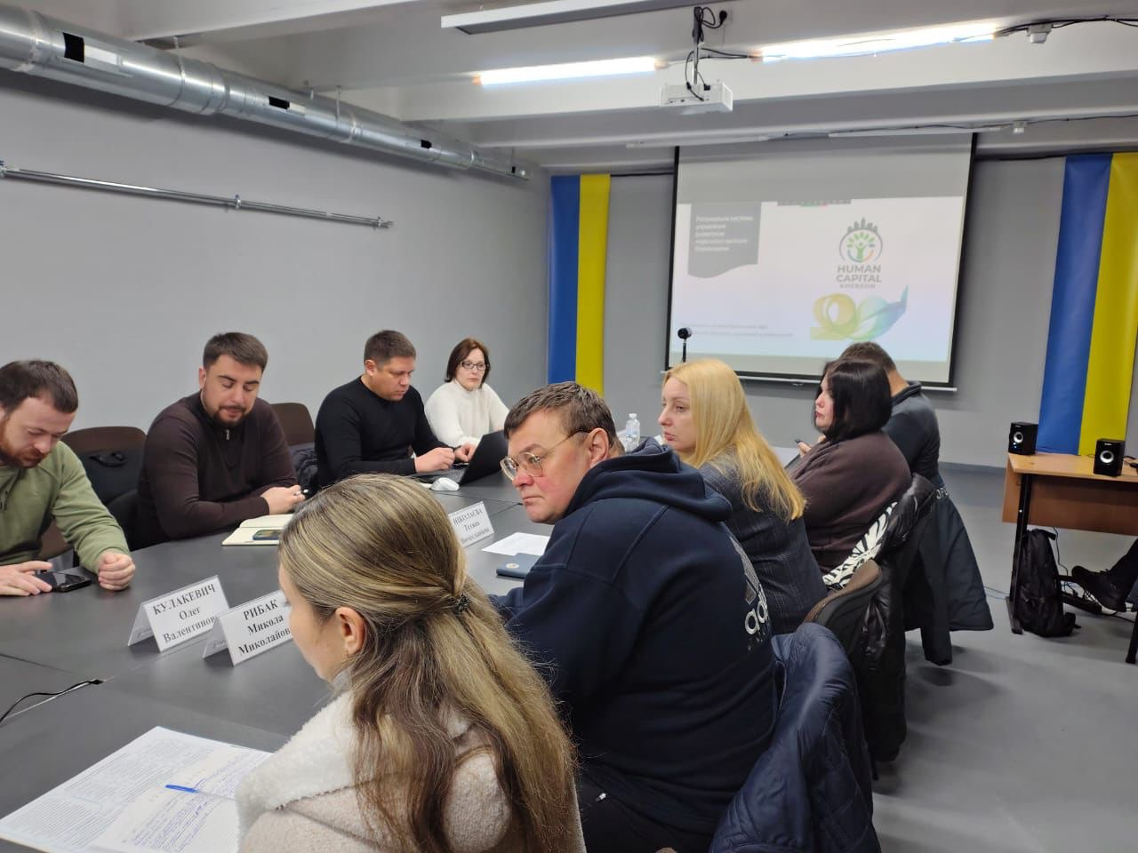 Учасники офіційної зустрічі сидять за столом у залі, декорованому українськими прапорами. На задньому плані розташований проєкційний екран із логотипом презентації.